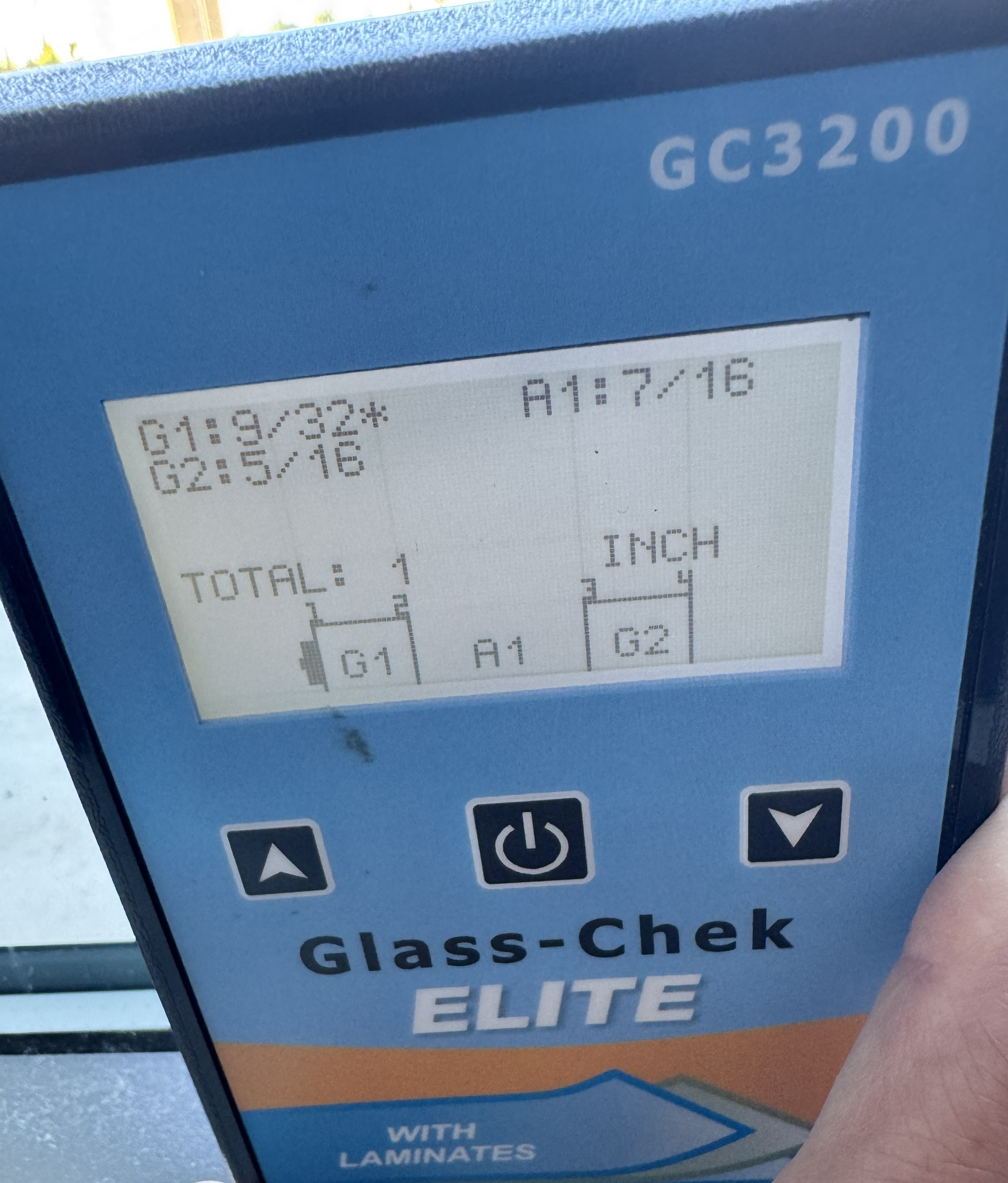Glass-Chek ELITE meter held against a double pane window, its display showing the thickness of each pane of glass and the air space between them.