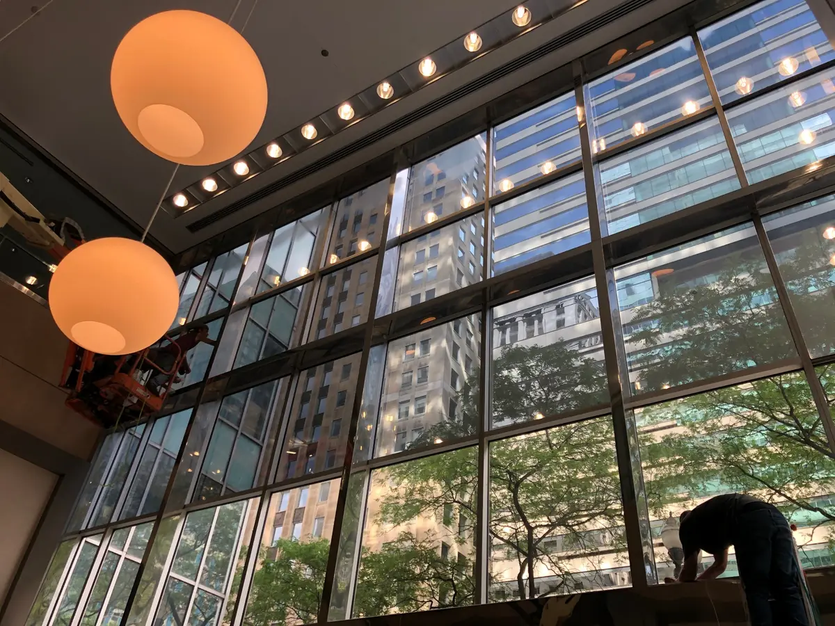 Two installers applying window film to a two-story glass curtain wall, one on a lift, inside an atrium with globe pendant lights.