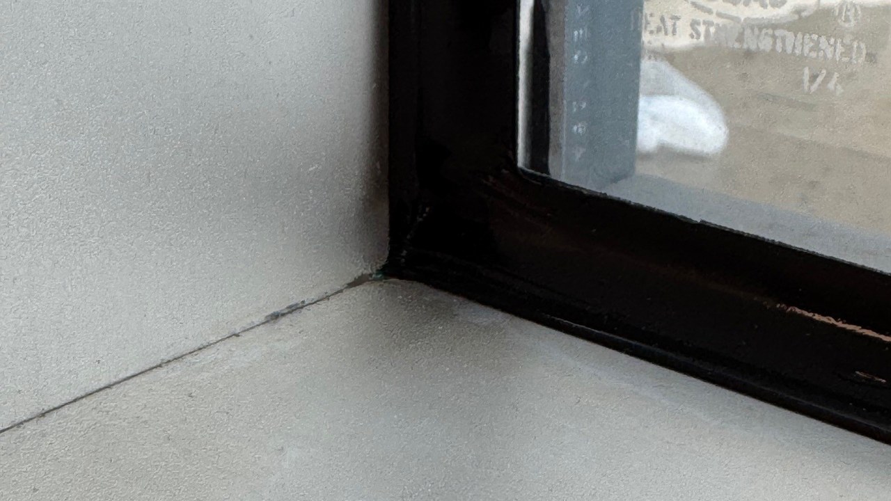 Close view of the bottom corner of a window from inside, with security window film on 1/4 inch heat strengthened glass and a black structural silicone bead along the side and bottom edges where the glass meets the frame.