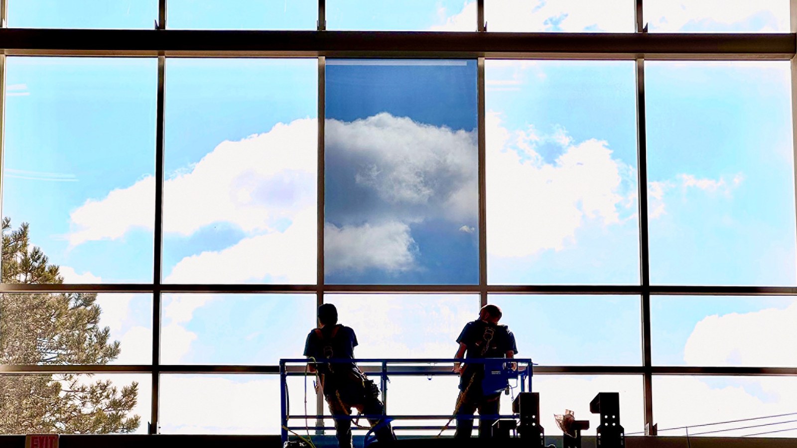 Two AAA Glass Tint installers on a scissor lift applying solar control window film to a tall commercial glass wall, with the first filmed pane showing reduced glare against the sky.