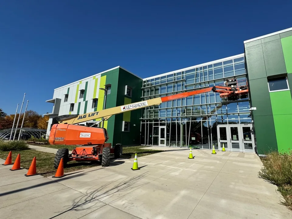 Boom lift at the glass entrance of a school with green wall panels, with safety cones on the walkway.